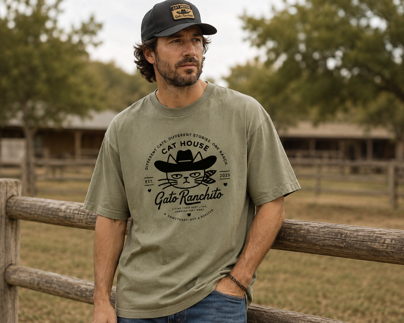 A man wearing an olive green graphic t-shirt with a black Cat House Gato Ranchito print, standing outdoors in front of a wooden fence and trees.