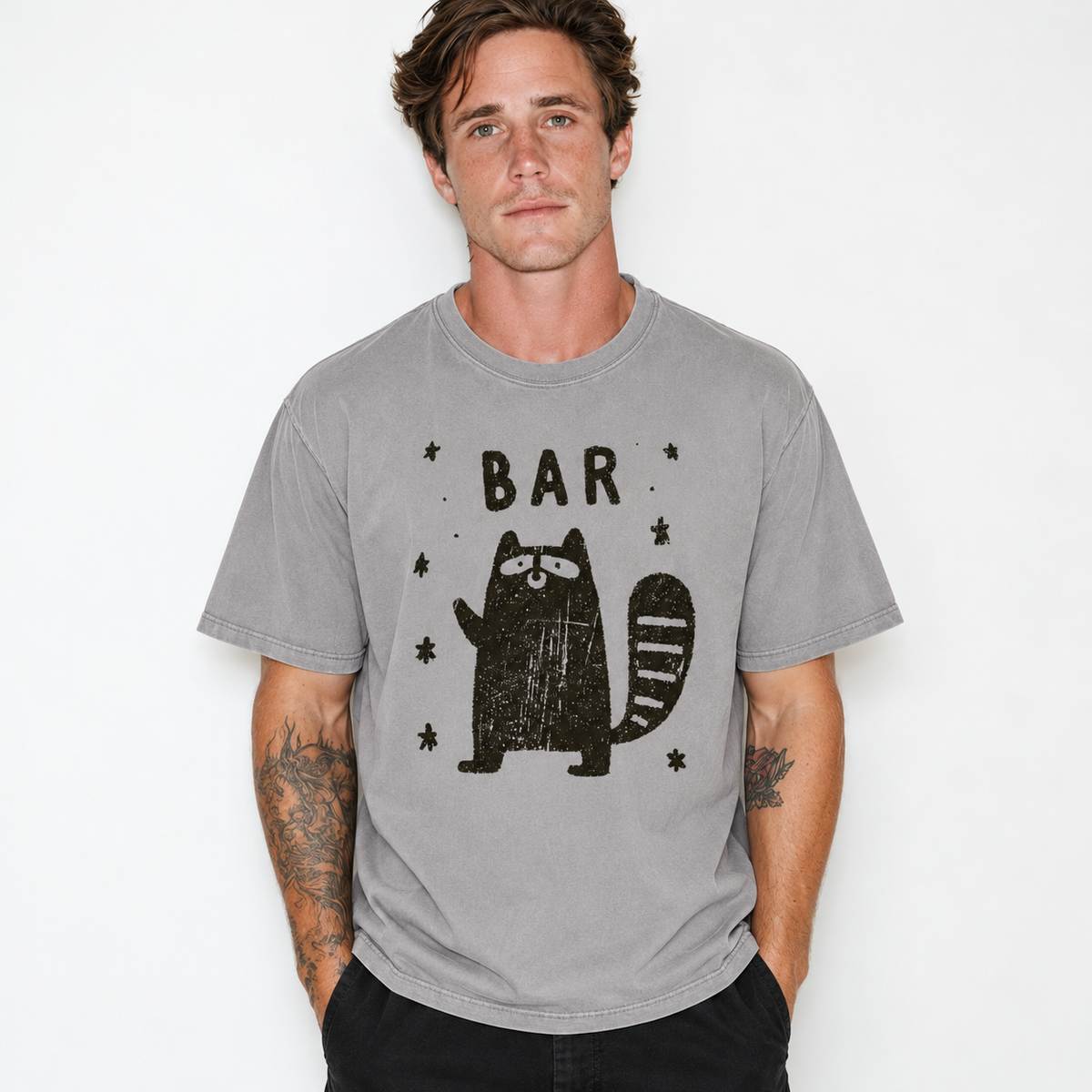 A man wearing a heather gray crewneck t-shirt with a distressed black graphic of a raccoon and the word 'BAR' on the front, set against a plain white background.