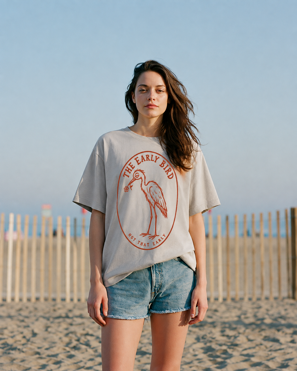 A woman stands on a sandy beach wearing an oversized light gray graphic T-shirt with a red bird illustration and the text 'THE EARLY BIRD BOSTON FEST' paired with denim shorts.