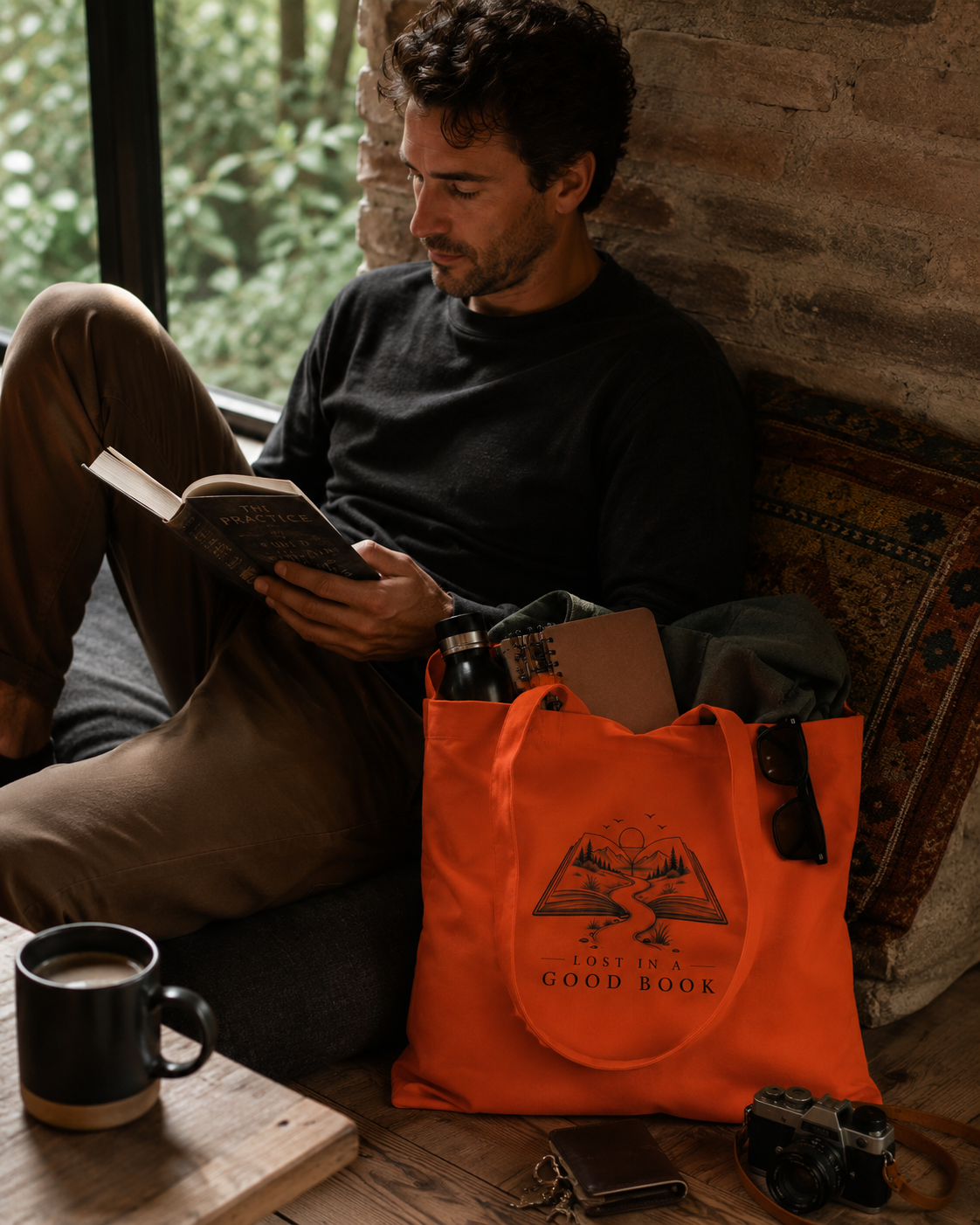 A man sits on a couch reading a book in a cozy room with a brick wall and window. An orange canvas tote bag with a black graphic and the text 'JUST ONE GOOD BOOK' sits on the floor next to him, along with a mug, camera, and other lifestyle props.