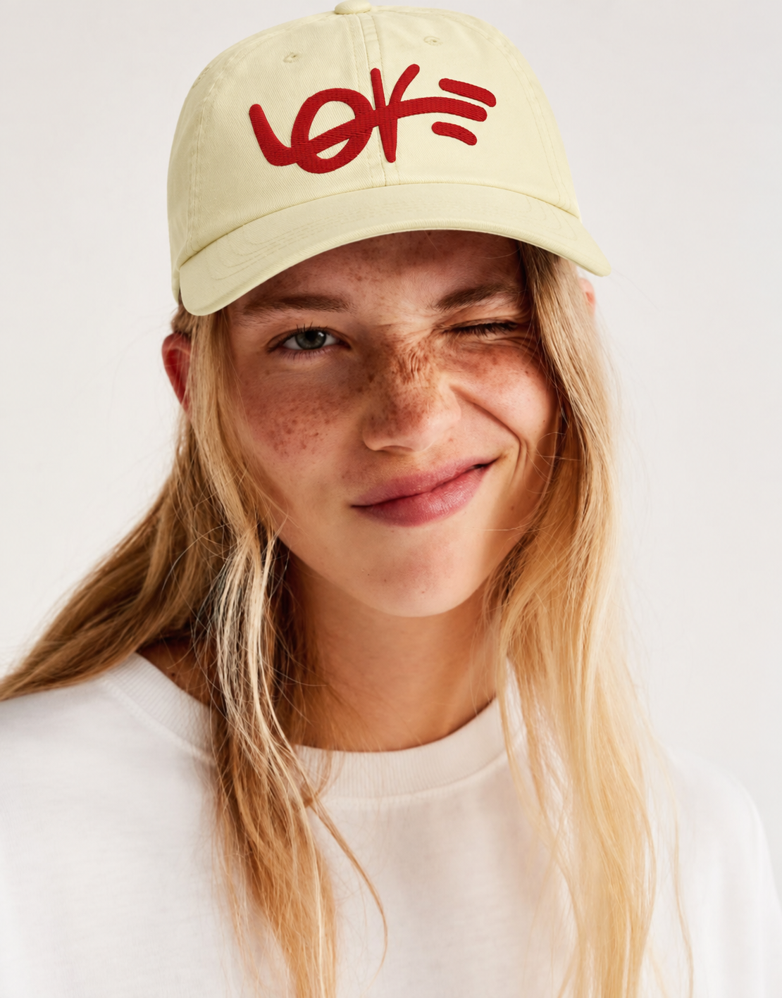 A close-up of a smiling woman with freckles wearing a cream-colored baseball cap with a red embroidered logo that reads 'LOVE'.