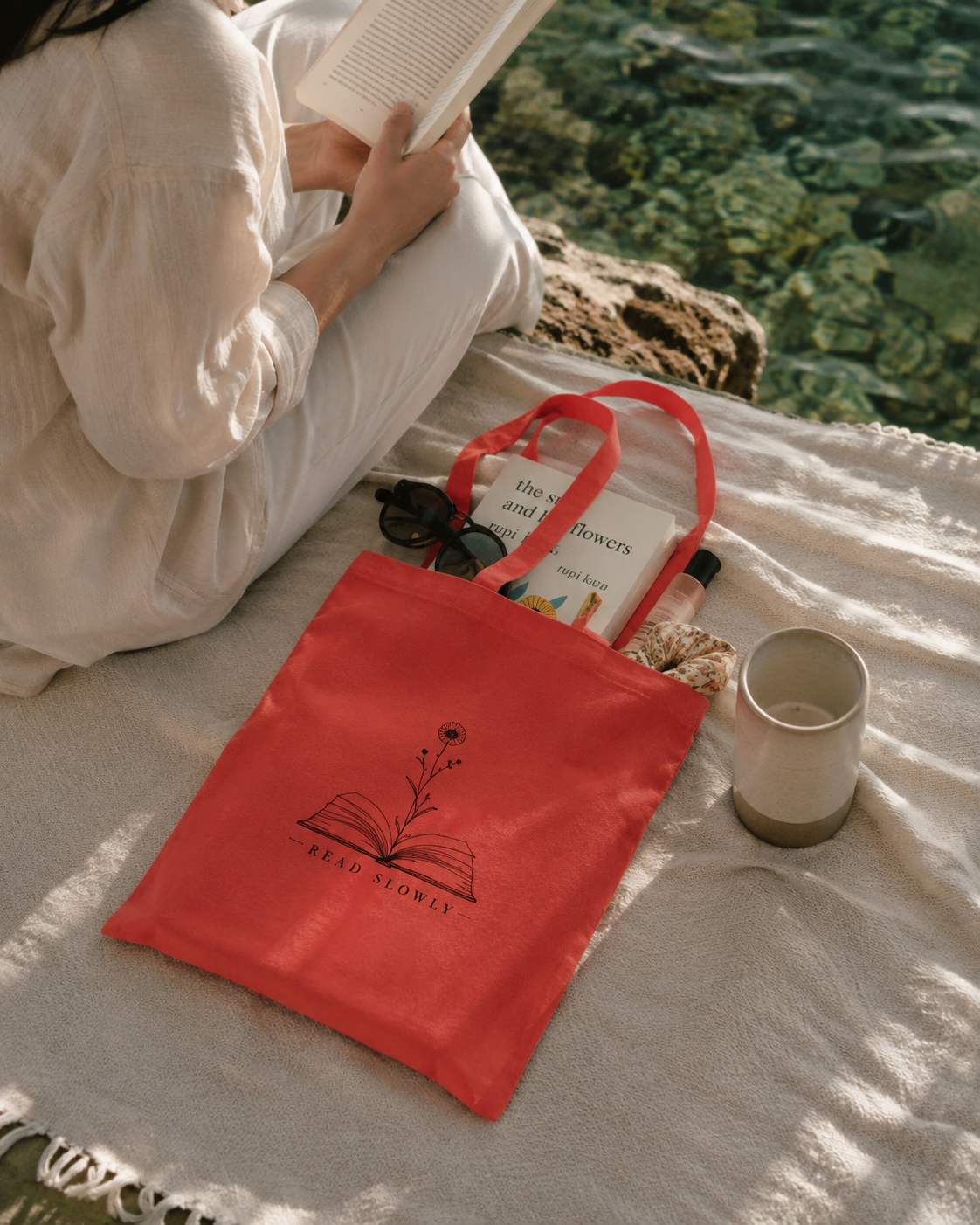 A red canvas tote bag with a black line-art graphic of an open book and flower, printed with the text 'REAL FLOWERS'. The bag is placed on a white blanket next to a person reading a book, with a cup nearby, set against a background of water and rocks.